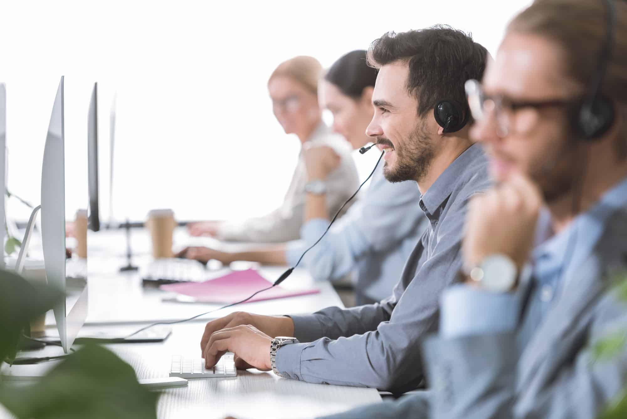 A row of call center phone operators sitting in a row, working at computers while wearing headsets.