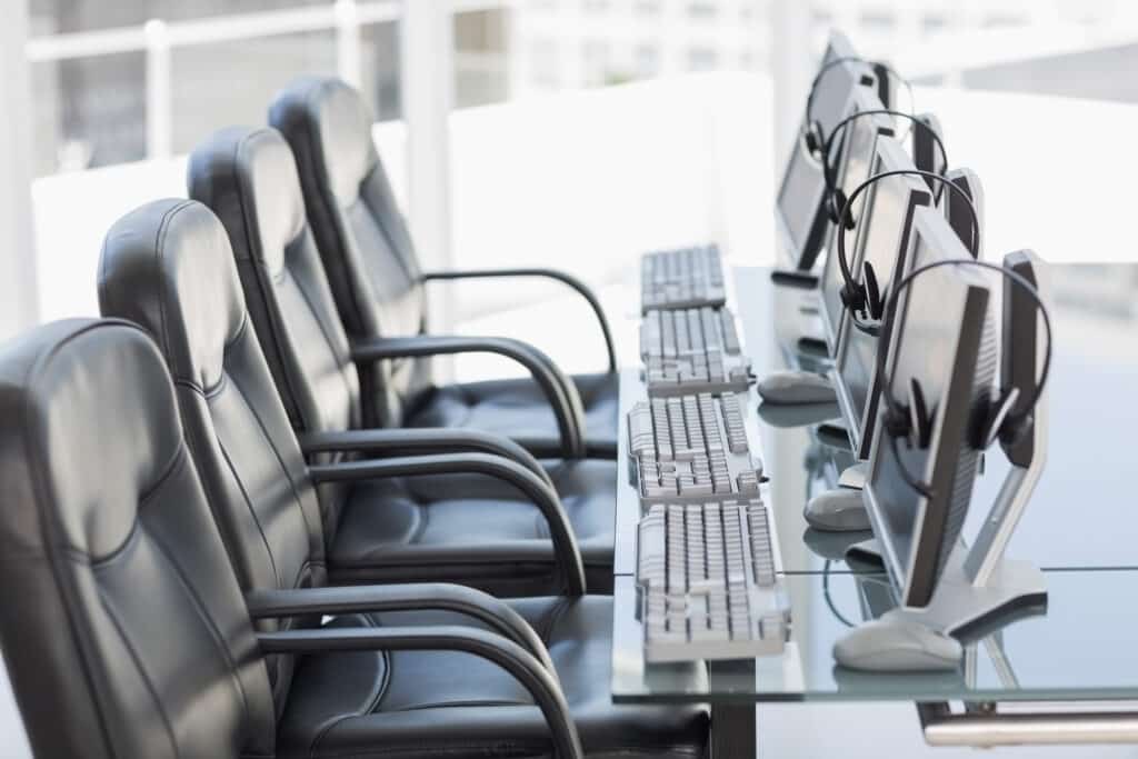 A row of empty leather office chairs lined up in front of desktop computers, each with headsets resting on the monitors.