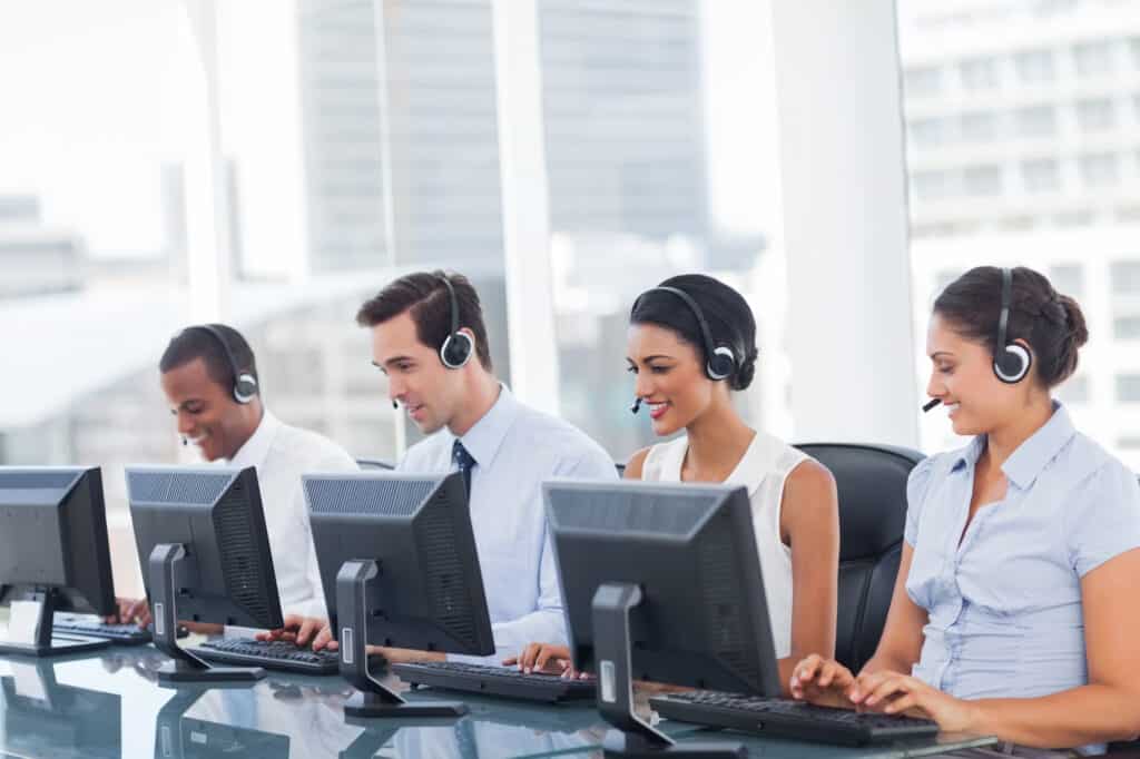 A row of call center representatives smile as they use their computers and speak to customers over their headsets.
