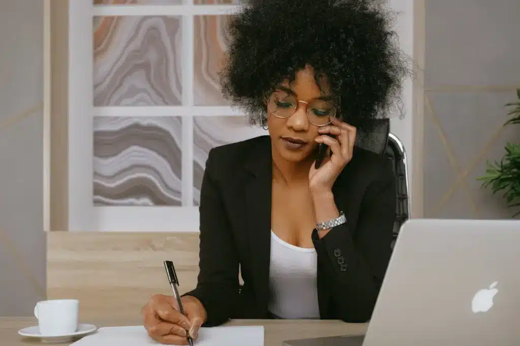 A woman in a black blazer is on the phone while she writes notes on a piece of paper with a pen; her call center AI solution is open on her laptop
