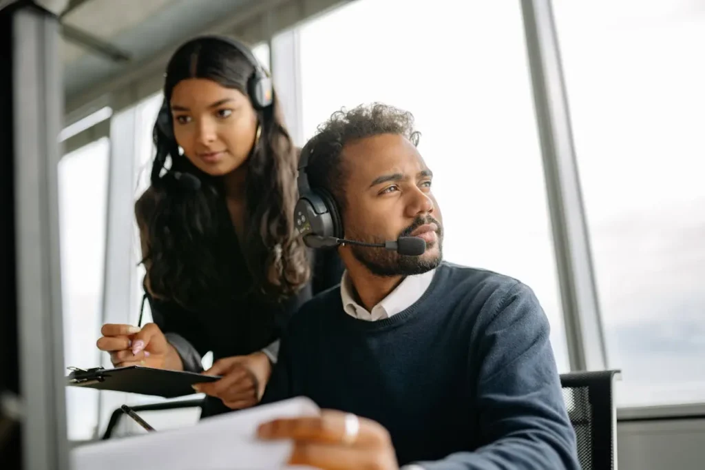 A call center agent refers to his AI solution to his left offscreen, while a female contact center agent takes notes on a clipboard on his right