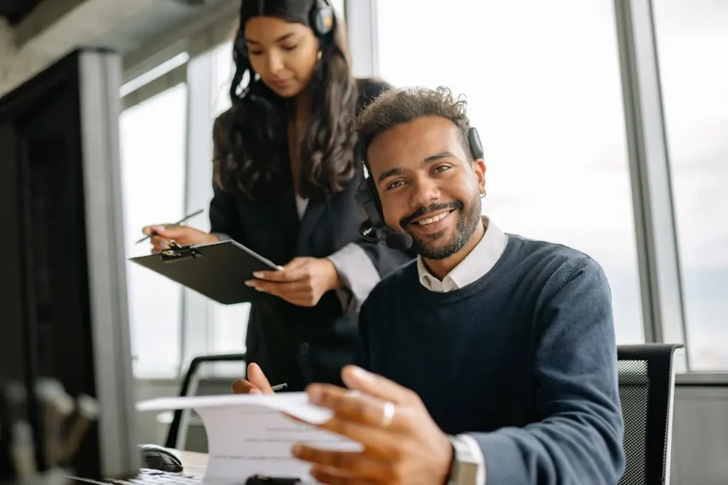 A smiling contact center agent looks at the camera, while another agent stands behind him with a clipboard