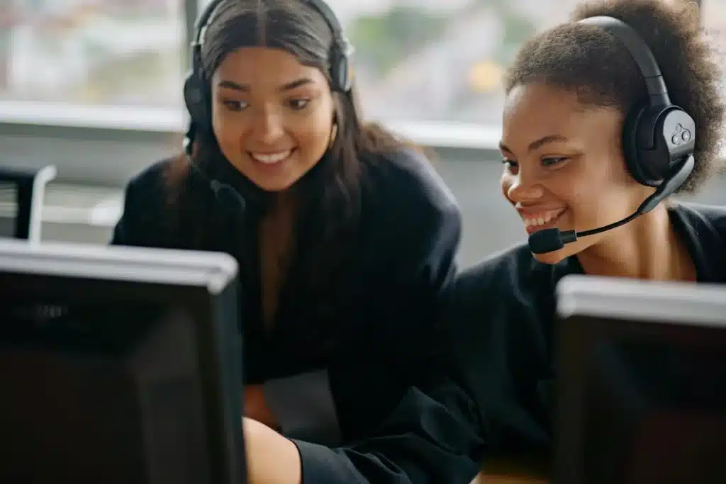 A contact center agent in a black blazer points out something on her monitor to another female contact center agent in a black blazer.